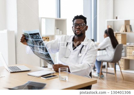 Doctor analyzing X-ray in medical office, showing healthcare, medical technology, and expertise. Image features male adult of African descent about 30s wearing professional attire in workspace Doctor analyzing X-ray in medical office, showing healthcare, medical technology, and expertise. Image features male adult of African descent about 30s wearing professional attire in workspace 130401306