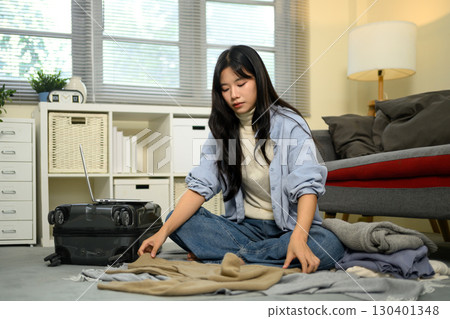 Female traveler organizing outfits on the floor next to suitcase before trip 130401348
