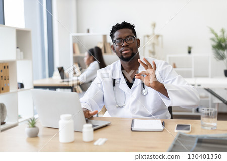 Young adult man doctor wearing white coat and glasses working on computer in bright office. Expression of confidence and professionalism, involved in healthcare and medicine, workplace 130401350