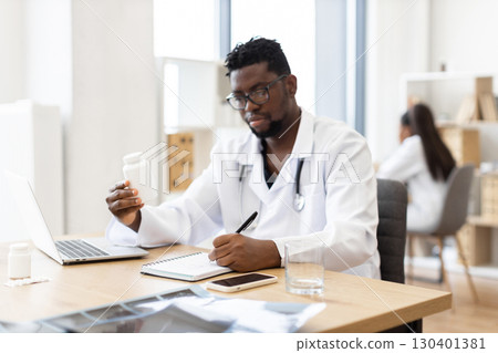 Male African doctor mid-30s sitting at desk writing prescription examining medication, office setting. Professional medical professional working healthcare tasks indoors. 130401381