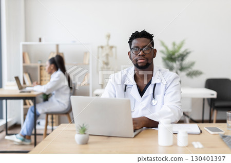 Professional young Black male doctor in white coat working at laptop in medical office. Working space includes professional tools, writing materials, and medical resources. 130401397