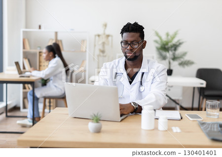 Male doctor using laptop in healthcare office while female colleague works in background. Modern medical environment with focus on technology, collaboration, and professional expertise. 130401401