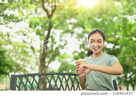 Smiling young woman holding a coffee cup and checking time on smartwatch in the park 130401437
