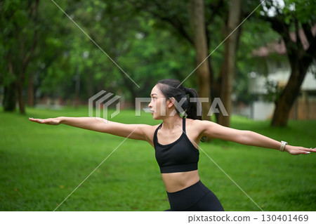 Young woman practicing yoga outdoors, performing warrior pose in a green park 130401469