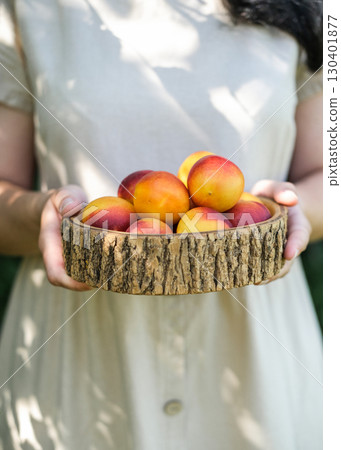 Woman in beige dress holds rustic wooden bowl filled with vibrant ripe nectarines, symbolizing summer harvest and fresh organic food. Ideal for food blogs, social media and healthy lifestyle content 130401877