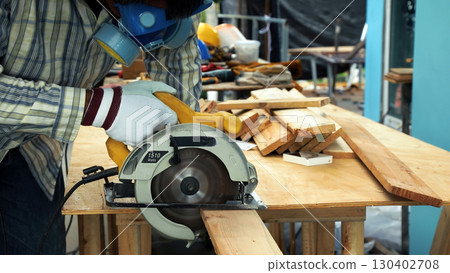 Carpenter using circular saw to cut wooden plank on outdoor workbench wearing gloves and protective mask Carpenter using circular saw to cut wooden plank on outdoor workbench wearing gloves and protective mask 130402708
