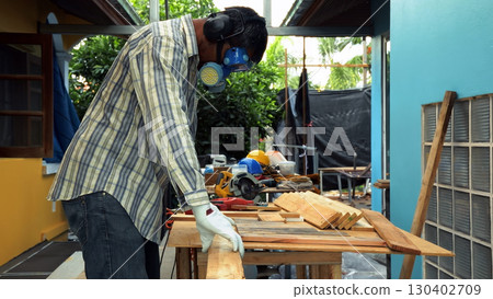 Carpenter lifting wooden plank on workbench while wearing gloves and safety gear during woodworking task Carpenter lifting wooden plank on workbench while wearing gloves and safety gear during woodworking task 130402709