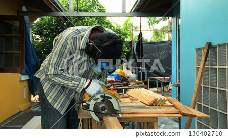 Carpenter using circular saw to cut wooden plank on workbench while wearing safety mask and gloves 130402710