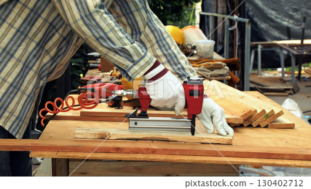 Close up of carpenter hand wearing gloves using red nail gun to fasten wooden plank on workbench in workshop 130402712