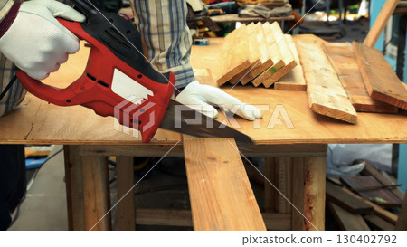 Worker cutting wooden plank with red electric saw on workbench wearing white gloves and striped shirt Worker cutting wooden plank with red electric saw on workbench wearing white gloves and striped shirt 130402792