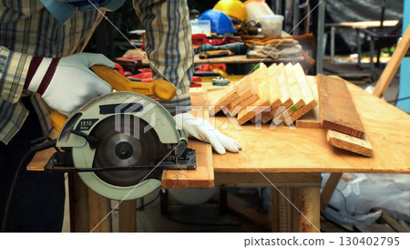 Carpenter cutting wooden plank with circular saw on workbench while wearing gloves and protective mask 130402795