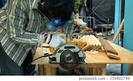 Carpenter cutting wooden board with circular saw on workbench while wearing safety mask gloves and protective gear 130402799