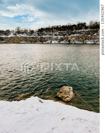 Snow covered shores of tranquil lake, featuring rugged rock formation and towering cliffs adorned with trees and snow, all beneath cloudy winter sky. Winter landscape with snow blanketed shoreline 130402967