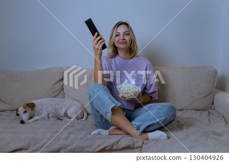 Home, evening: Young woman in casual clothes sits cross-legged on a sofa, holding popcorn and a remote, watching TV with a soft smile. A small dog sleeps peacefully next to her. 130404926