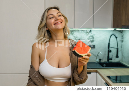 A sensual woman enjoys a slice of juicy watermelon in a modern kitchen. Confident, playful, and radiant, she savors the moment with a hint of summer flirtation. 130404930