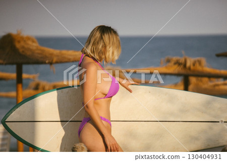 Attractive young woman in a purple bikini posing with a surfboard on the beach, enjoying summer by the sea, confident and relaxed, ocean waves in the background. 130404931
