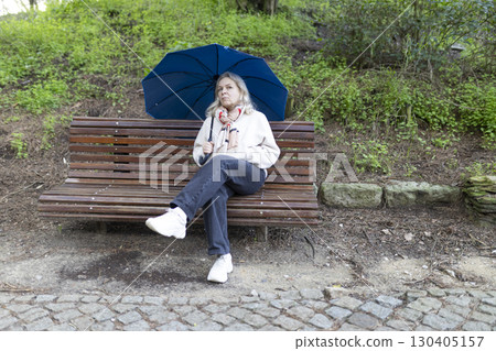 Senior woman holding umbrella sitting on bench in park after rain 130405157