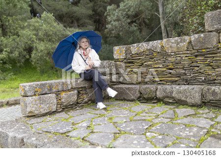 Senior woman holding blue umbrella sitting on stone wall in park Senior woman holding blue umbrella sitting on stone wall in park 130405168