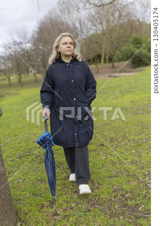 Senior woman walking in park with closed umbrella on cloudy day Senior woman walking in park with closed umbrella on cloudy day 130405174