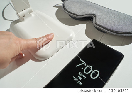 Hand turning off alarm on smartphone with white clock and eye mask in morning 130405221