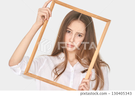 Young caucasian female in white shirt holding wooden frame against plain 130405343