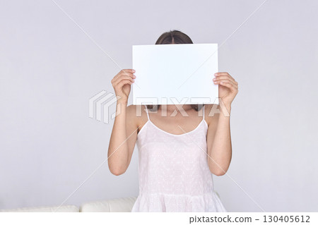 Female holding blank paper in front of face wearing white dress against plain 130405612