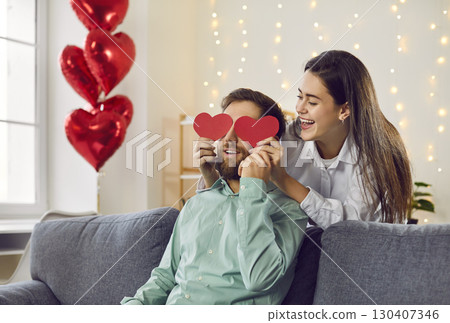 Happy young woman covering eyes of her boyfriend with red paper hearts Happy young woman covering eyes of her boyfriend with red paper hearts 130407346