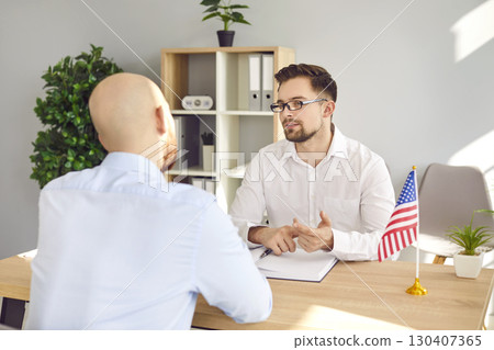 Consul sitting at desk with young man and talking to him about visa and immigration to USA 130407365
