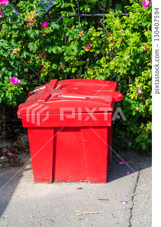 A Vibrant Red Trash Bin Beautifully Surrounded by Lush Green Foliage A Vibrant Red Trash Bin Beautifully Surrounded by Lush Green Foliage 130407594