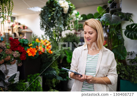 Florist managing her shop with a tablet, surrounded by plants and flowers Florist managing her shop with a tablet, surrounded by plants and flowers 130408727
