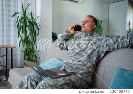 Happy Soldier Relaxing on Sofa, Talking on Phone and Using Laptop 130408773