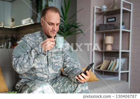 Soldier relaxing on sofa, drinking coffee and using smartphone Soldier relaxing on sofa, drinking coffee and using smartphone 130408787