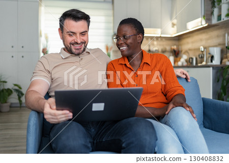 Happy multi-ethnic couple using laptop at home, relaxing on sofa 130408832
