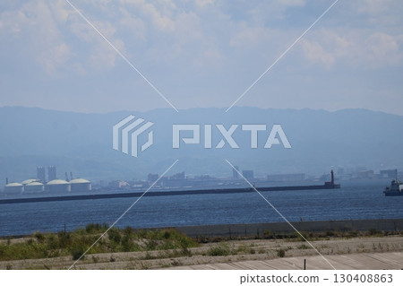 A jetty with a lighthouse at the end of the embankment, a ship, the blue ocean, the industrial complex, the hazy cityscape, the mountains, and the cloudy sky. 130408863