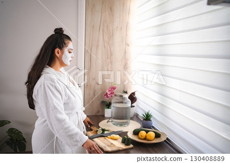 Young woman wearing bathrobe and face mask preparing detox water with lemon and cucumber in the kitchen 130408889