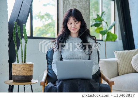 Portrait of young woman sitting on chair typing on laptop, in home interior 130408937