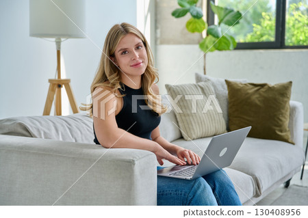 Portrait of young woman sitting on sofa with laptop, in home interior 130408956