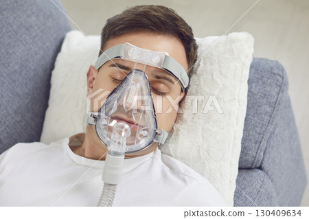 A young man with an oxygen mask on his face sleeps on the sofa at home 130409634