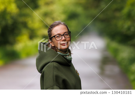 Woman in green hoodie stands on a serene path surrounded by lush greenery on a cloudy day Woman in green hoodie stands on a serene path surrounded by lush greenery on a cloudy day 130410181