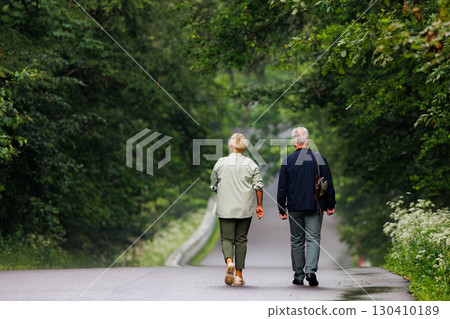 Elderly couple walking hand in hand down a serene forest path surrounded by lush greenery 130410189