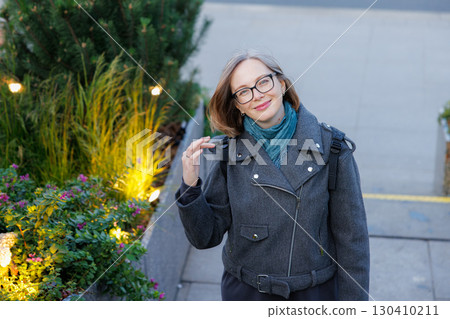 Woman in stylish coat stands by plants and smiles in urban setting during daylight Woman in stylish coat stands by plants and smiles in urban setting during daylight 130410211