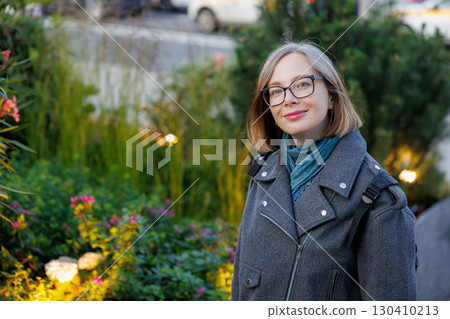 Smiling woman poses in a green urban garden on a sunny day while wearing stylish glasses and a warm jacket Smiling woman poses in a green urban garden on a sunny day while wearing stylish glasses and a warm jacket 130410213