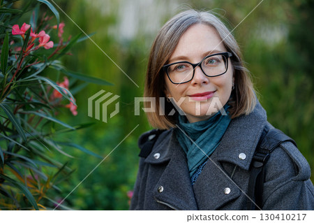 Woman in a gray coat enjoys a blooming garden with pink flowers in a sunny environment during autumn Woman in a gray coat enjoys a blooming garden with pink flowers in a sunny environment during autumn 130410217