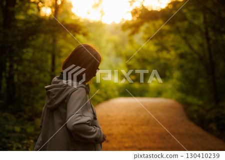 Woman walking on a forest path during sunset in a lush green setting with golden light illuminating the scene 130410239