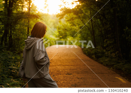 Woman enjoys a peaceful evening stroll along a sunlit forest path in golden hour glow Woman enjoys a peaceful evening stroll along a sunlit forest path in golden hour glow 130410240