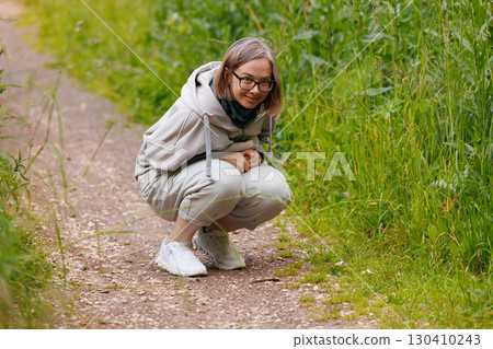 Person crouching on a nature path observing plants in a lush green environment during daylight hours Person crouching on a nature path observing plants in a lush green environment during daylight hours 130410243