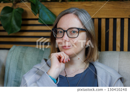 Woman with glasses sitting indoors, thoughtfully smiling while resting her chin on her hand Woman with glasses sitting indoors, thoughtfully smiling while resting her chin on her hand 130410248