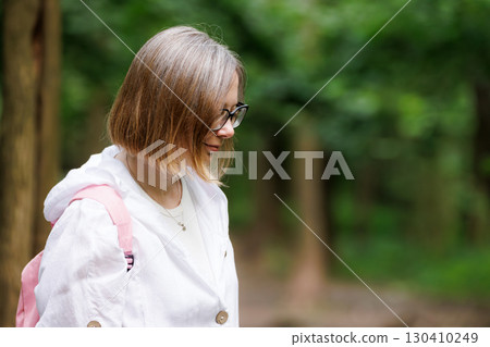Young woman with glasses walks through a lush green forest on a sunny day while wearing a backpack 130410249