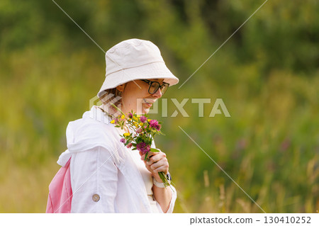 Young woman with glasses collects wildflowers in a sunny field during a warm afternoon 130410252