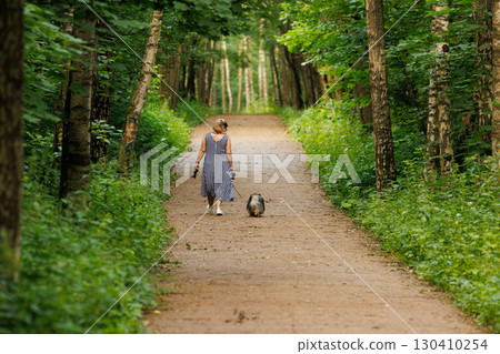 Woman walks pet dog along a peaceful forest path surrounded by lush green trees in summer Woman walks pet dog along a peaceful forest path surrounded by lush green trees in summer 130410254